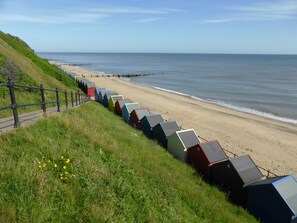 A beach with colorful beach huts aligned along the shore, a grassy area with a fence, and the ocean in the background.