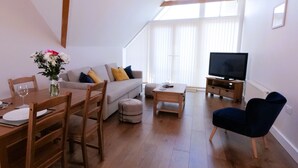 Un salón amplio con piso de madera, un conjunto de sofás, una mesa de comedor con sillas, un televisor y un ventanal que permite la entrada de luz natural.