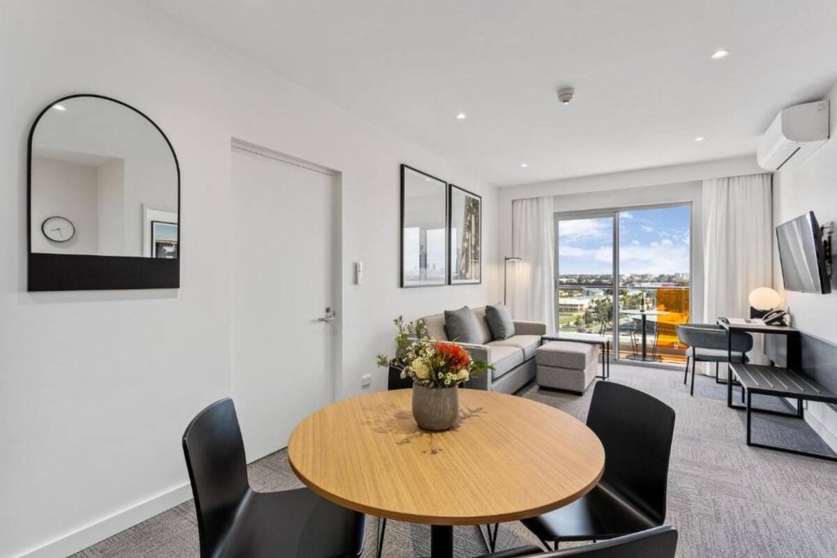 A modern hotel room with a round dining table, black chairs, a sofa, and a large window offering a city view.