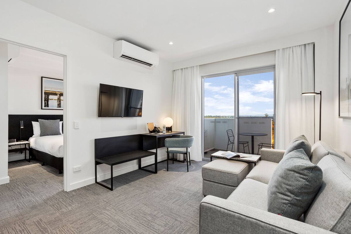 A modern hotel room with a bed, a sofa, a desk, and a balcony with a view.