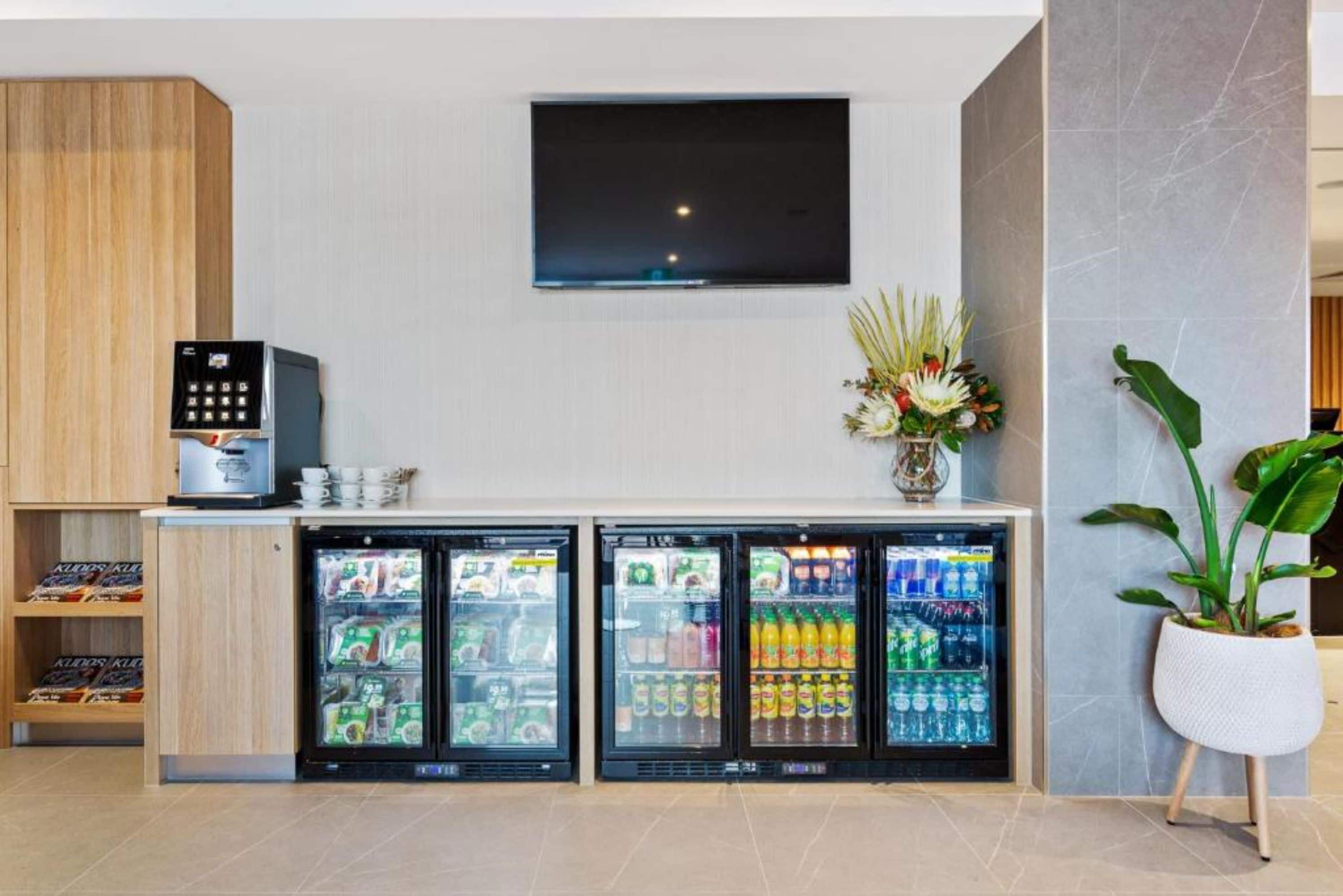 Un vestíbulo de hotel con una máquina de café, tres refrigeradores para bebidas, un televisor de pantalla plana y una planta en maceta.