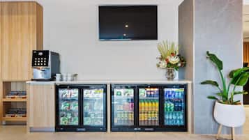 Un vestíbulo de hotel con una máquina de café, tres refrigeradores para bebidas, un televisor de pantalla plana y una planta en maceta.