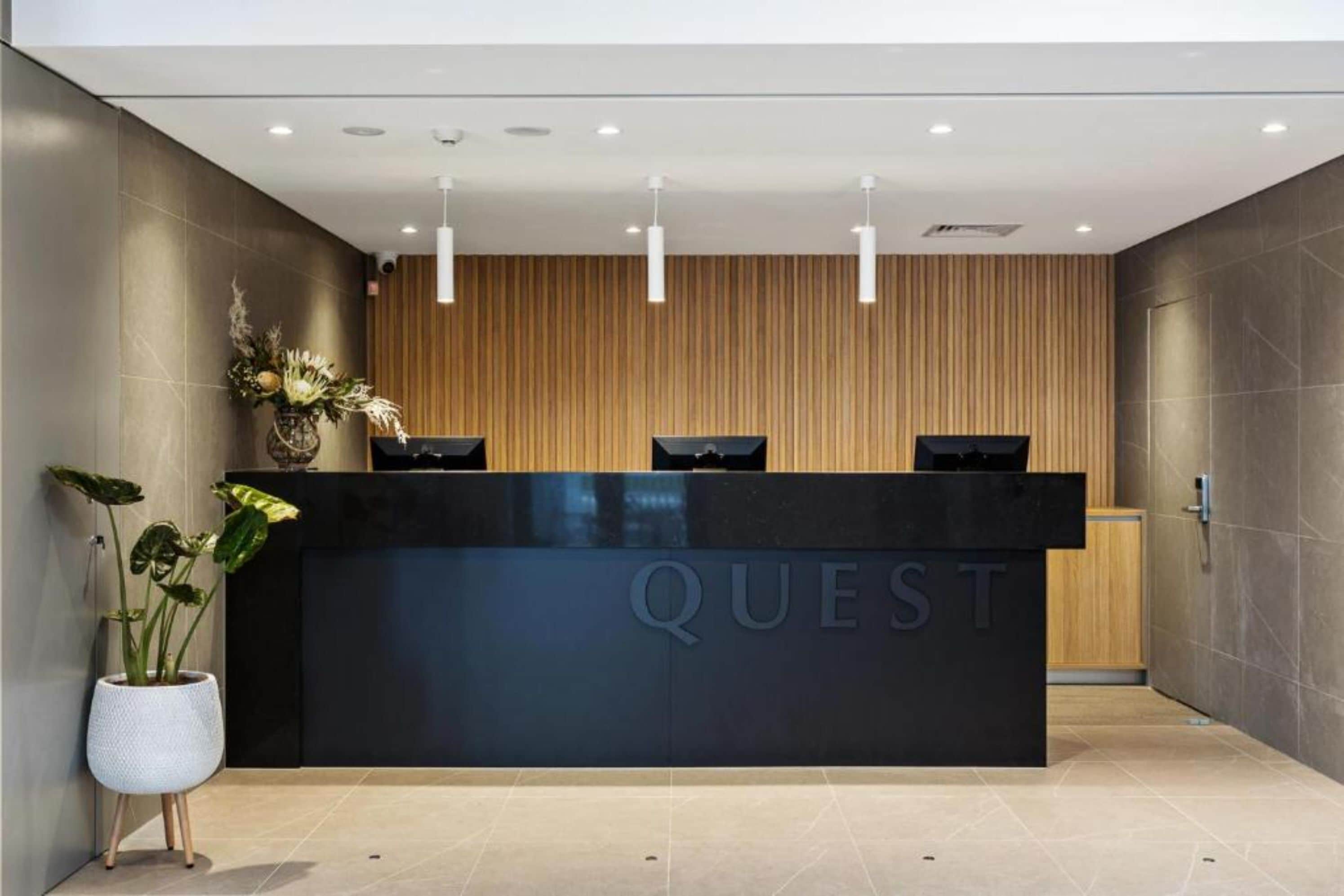 A modern reception desk with the word "QUEST" on it, flanked by wooden paneling and a potted plant.