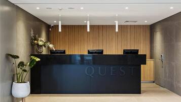 A modern reception desk with the word "QUEST" on it, flanked by wooden paneling and a potted plant.