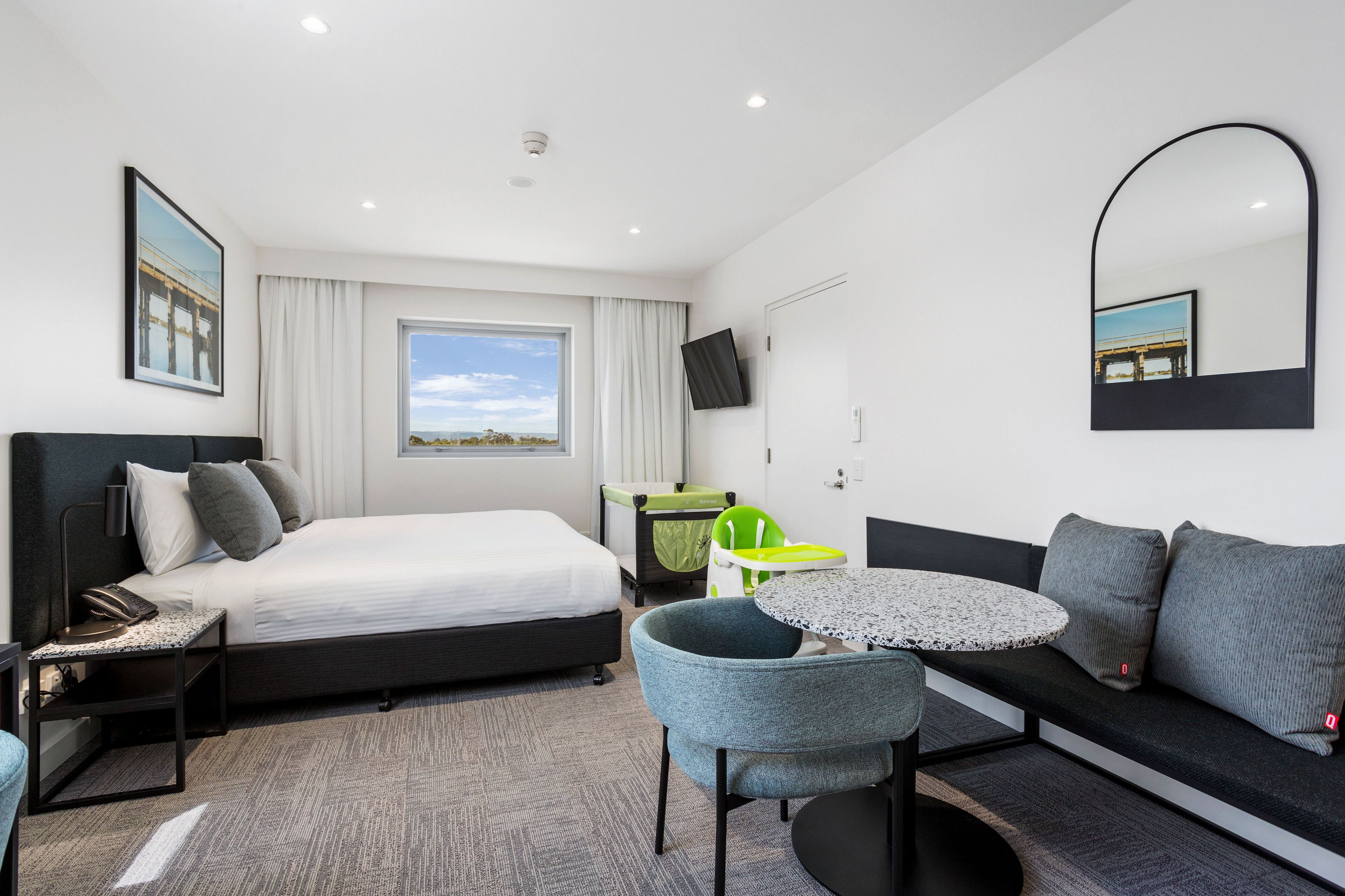 Et moderne hotelværelse med seng, sofa, lille bord, stol, skrivebord, fjernsyn og et vindue med udsigt til en bro.