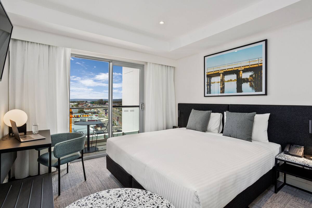 A modern hotel room with a large bed, a desk with a laptop, a chair, a round table, and a balcony with a city view.