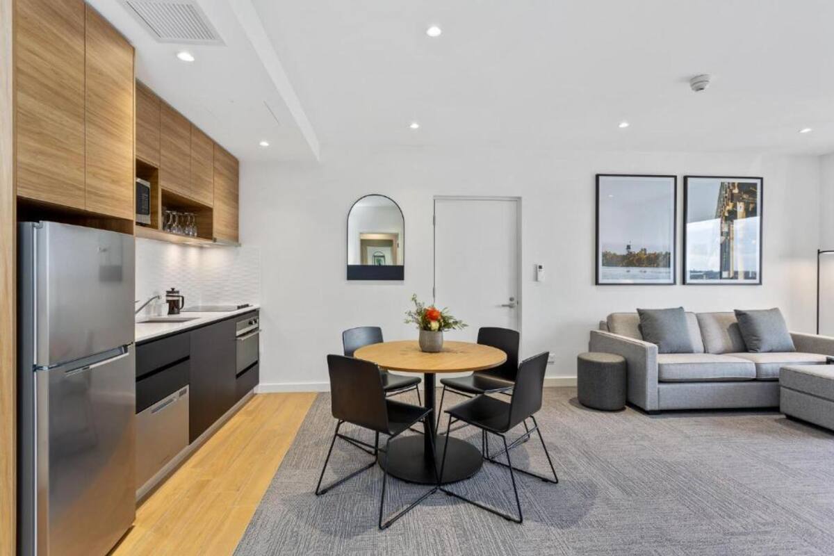 A modern kitchen with a built-in refrigerator, wooden cabinets, and a round dining table with chairs. There is a grey sofa and a small round table in the living area.