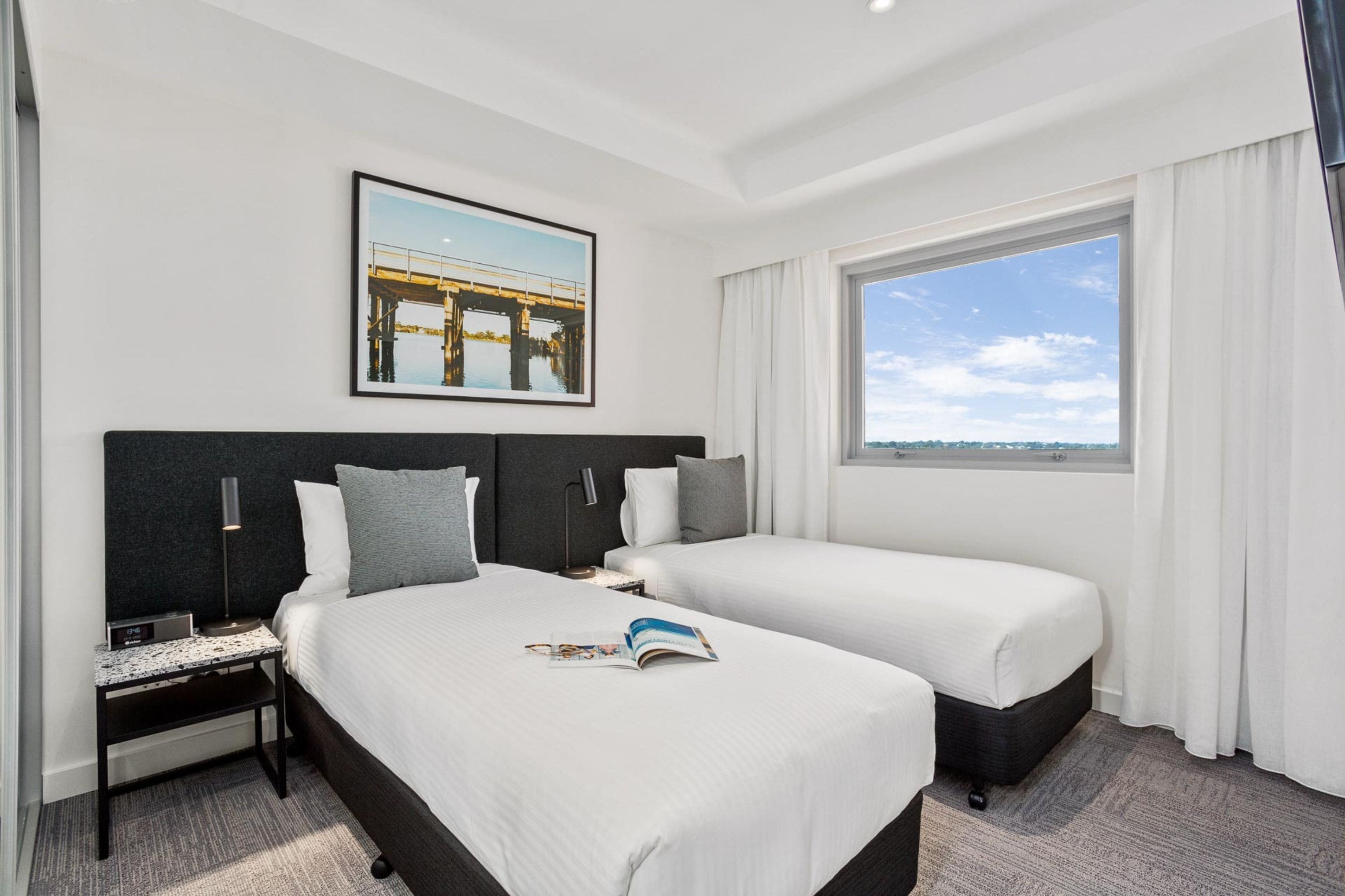 Habitación de hotel con dos camas, una mesita de noche, una lámpara, un cuadro en la pared y una ventana con vistas.