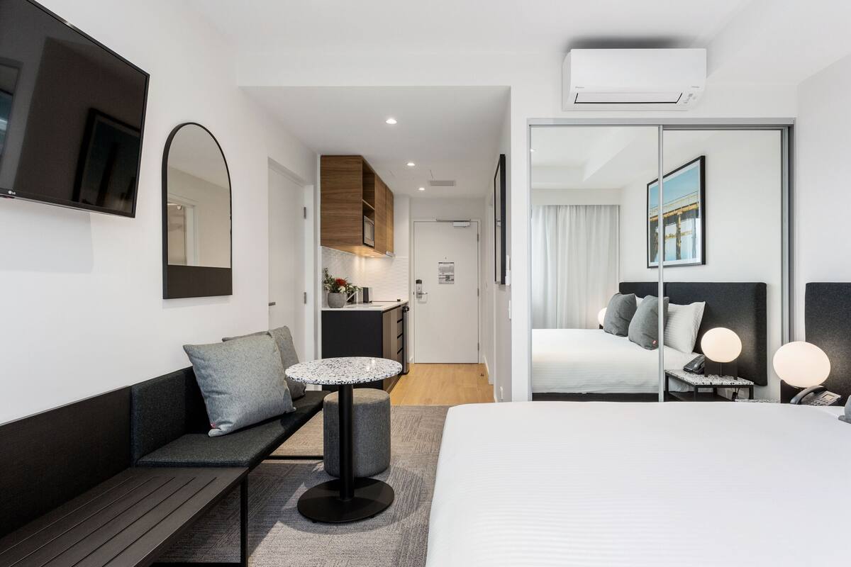A modern hotel room with a bed, a small round table, a sofa, a desk, and a kitchen area in the background.