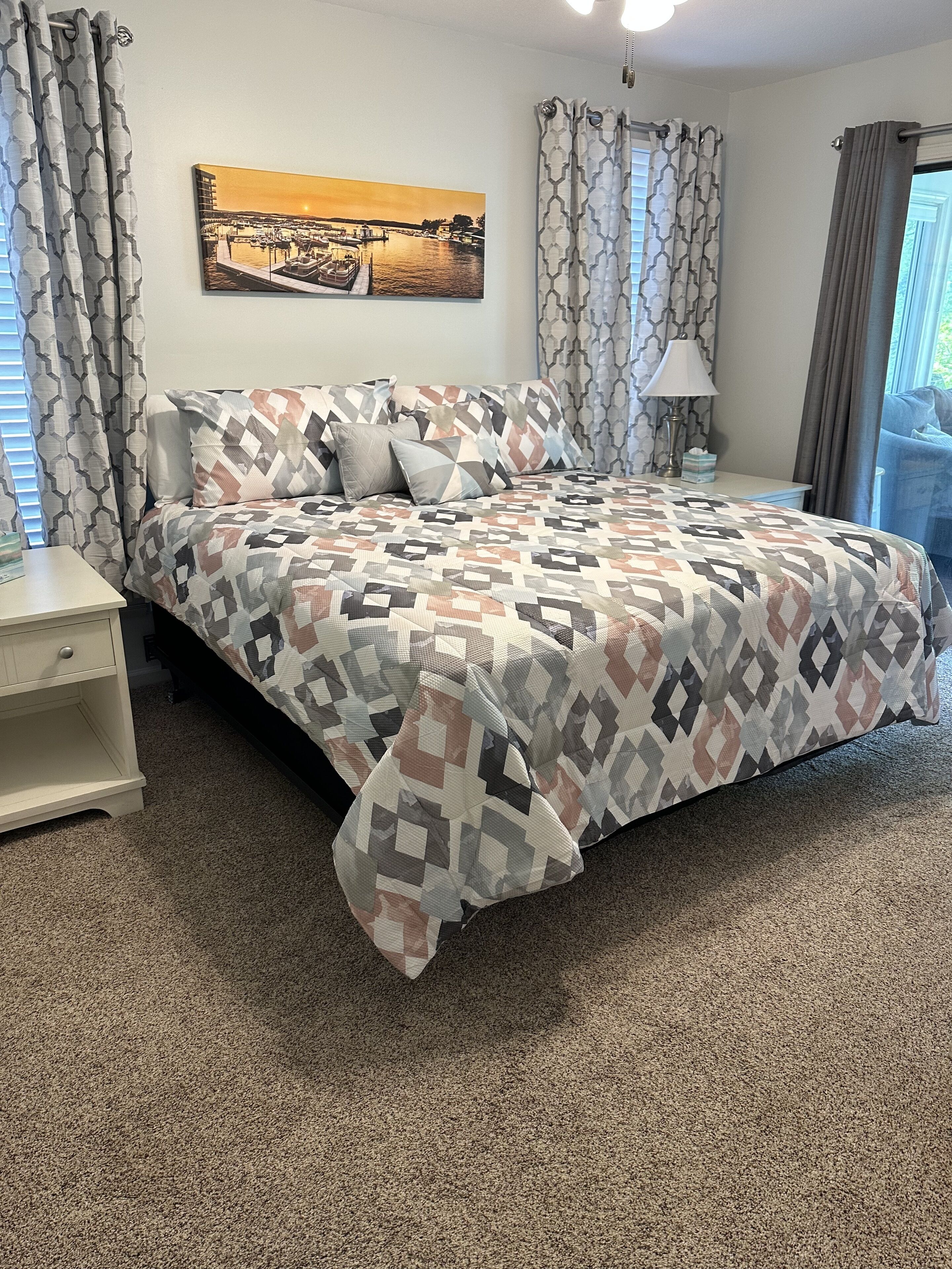 Une chambre à coucher avec une couvre-lit à motifs, une table de chevet, une lampe et une fenêtre avec des rideaux.
