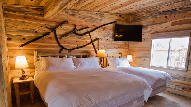 A wooden cabin bedroom with a large bed, two bedside tables with lamps, and a TV mounted on the wall. - Tack Room Studio on 280 acre Sevier River Ranch - 1 King and 2 Twin Beds (Hatch)
