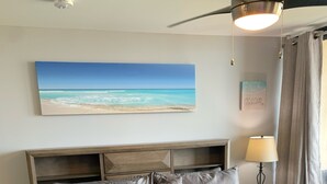 Een net opgemaakt bed met een grijze deken en kussens, een plafondventilator, een wandlamp en een schilderij met een strandthema erboven.