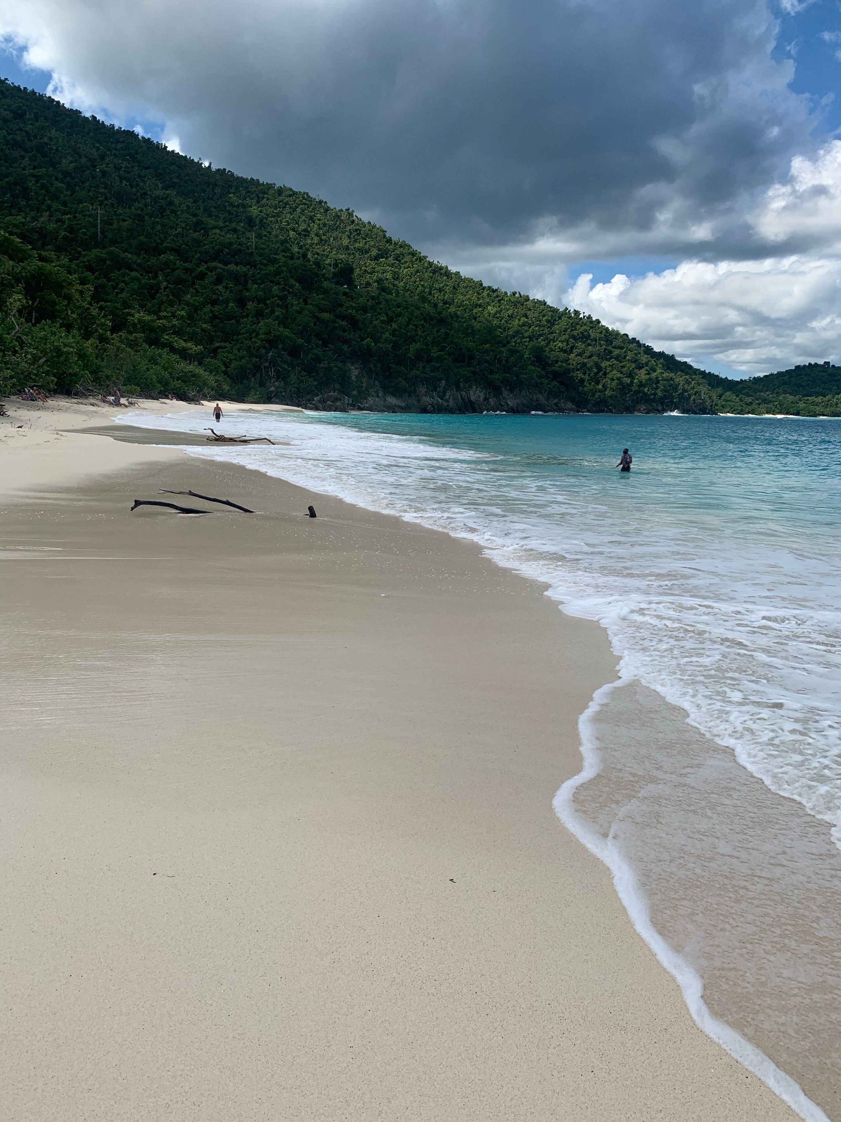 A beach with clear turquoise water, a sandy shore, and a forested hill in the background.