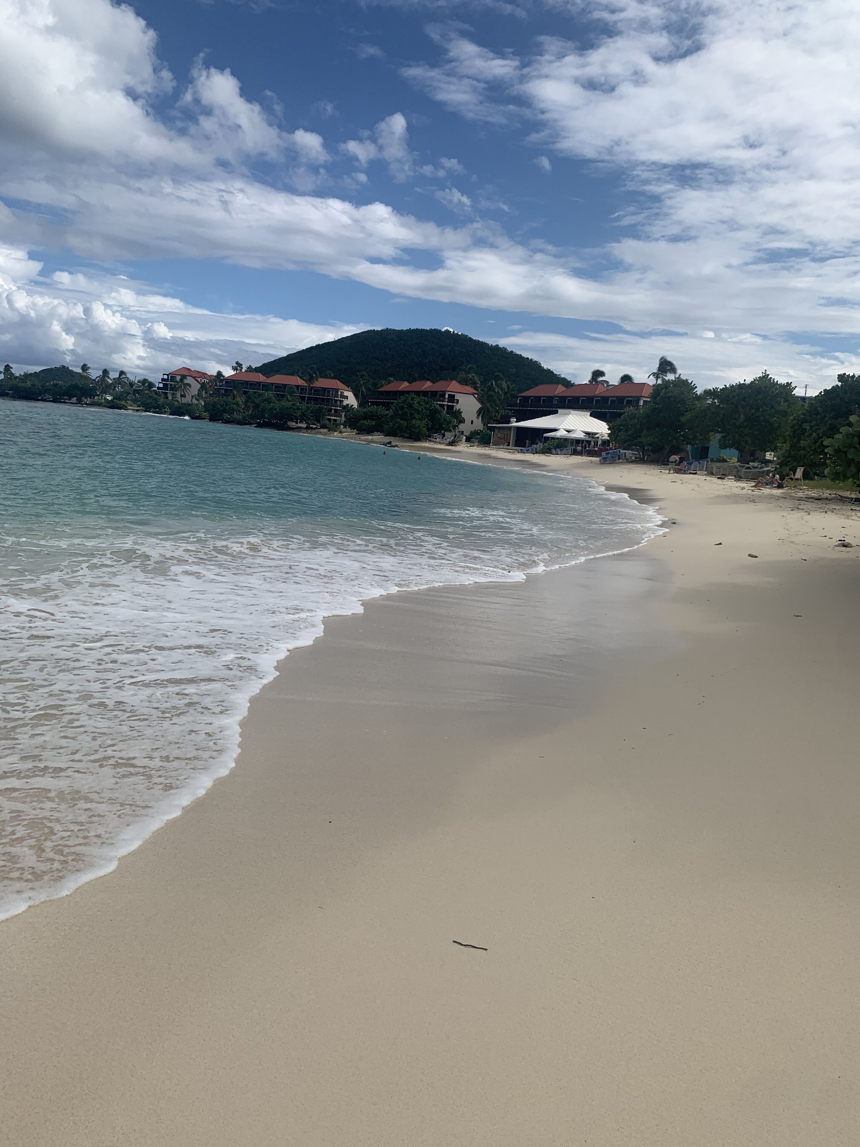 A beach with a sandy shore, a calm sea, and buildings with red roofs in the background.