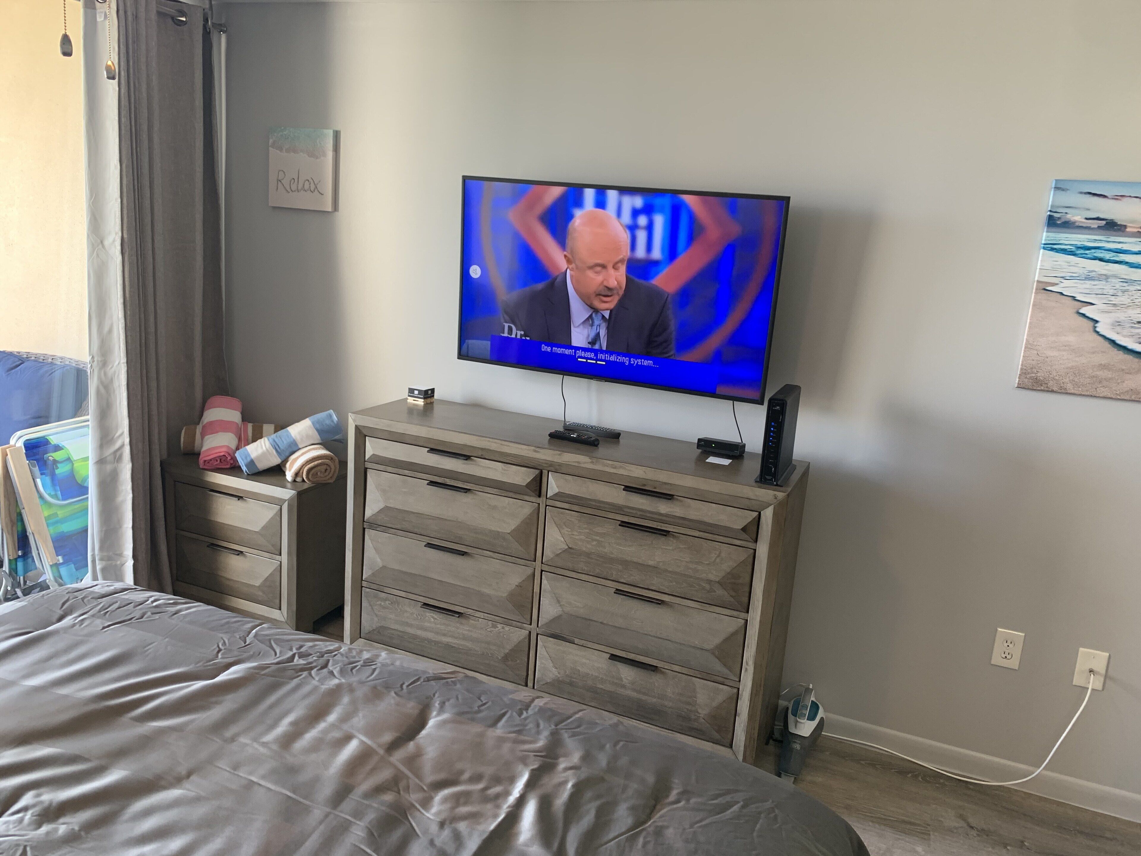 Quarto com cama, televisão em cômoda de madeira, quadro na parede e janela com cortinas.