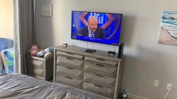 Quarto com cama, televisão em cômoda de madeira, quadro na parede e janela com cortinas.