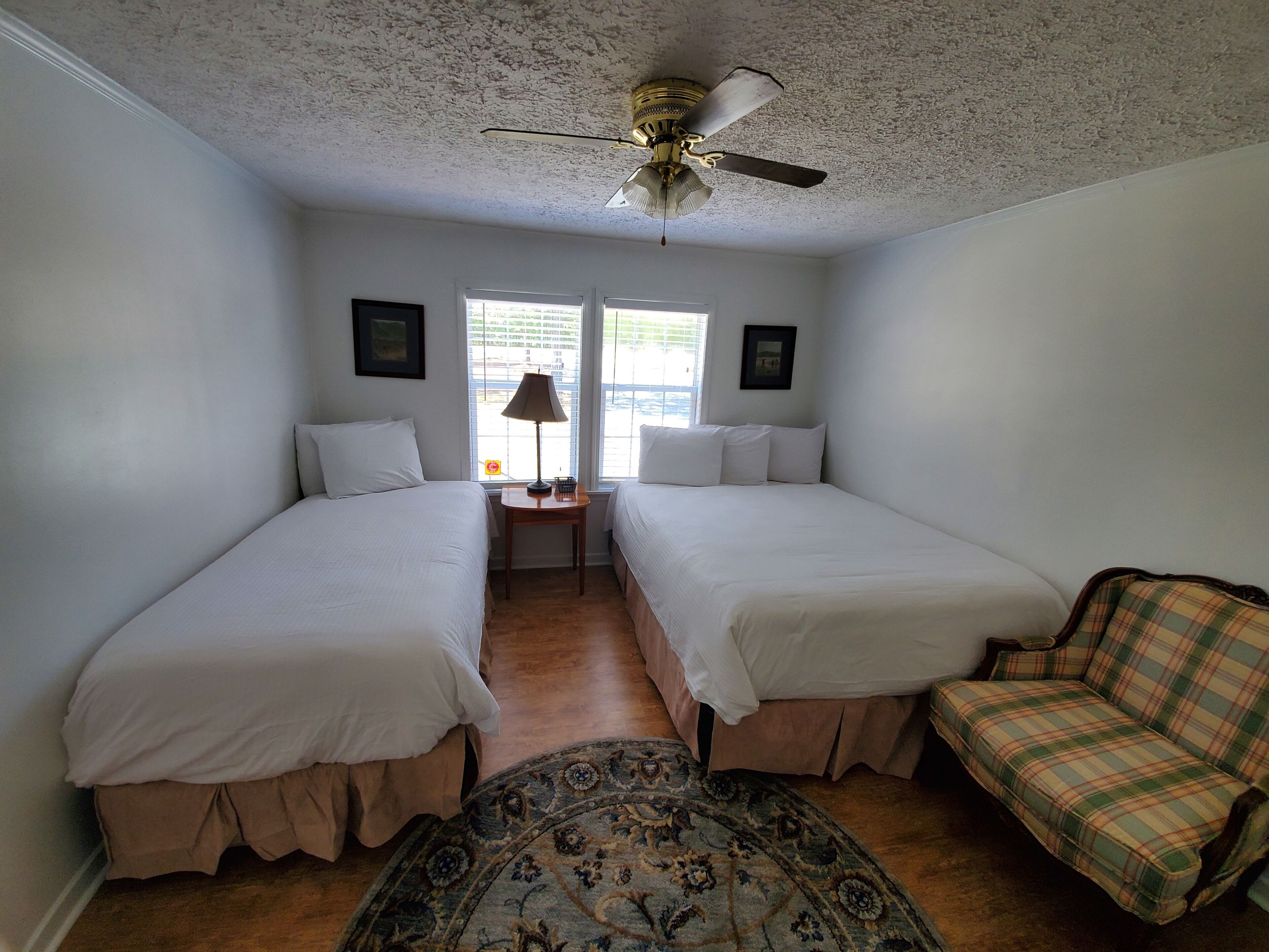 A room with two single beds, a small table with a lamp, a chair, and a ceiling fan.