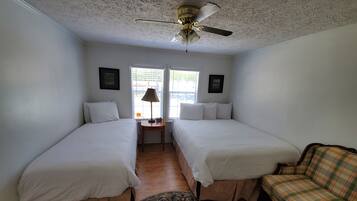 A room with two single beds, a small table with a lamp, a chair, and a ceiling fan.
