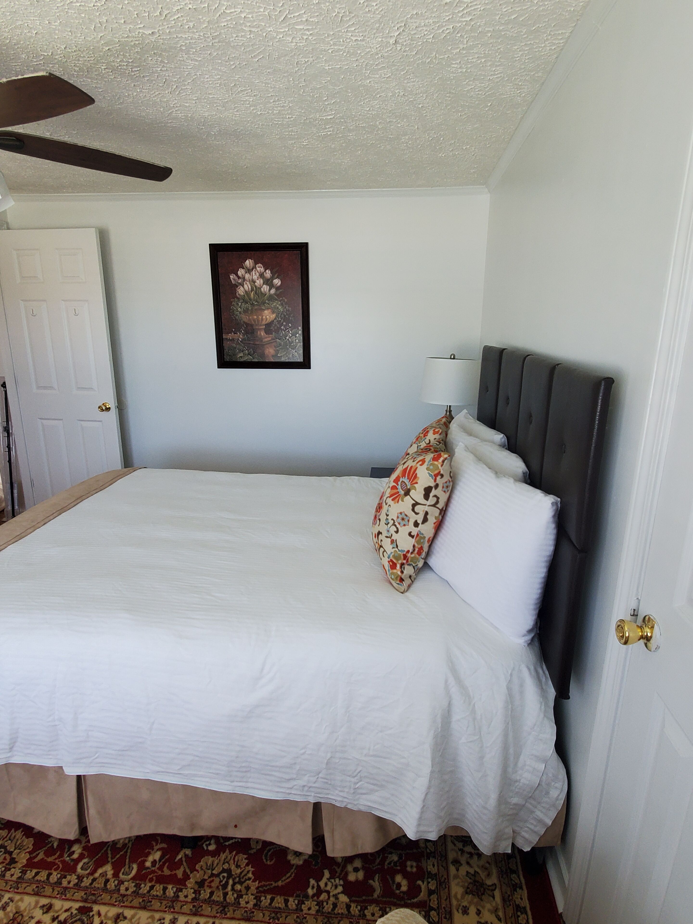 Une chambre à coucher avec un grand lit, une tête de lit, une porte, un ventilateur de plafond et un tableau encadré au mur.