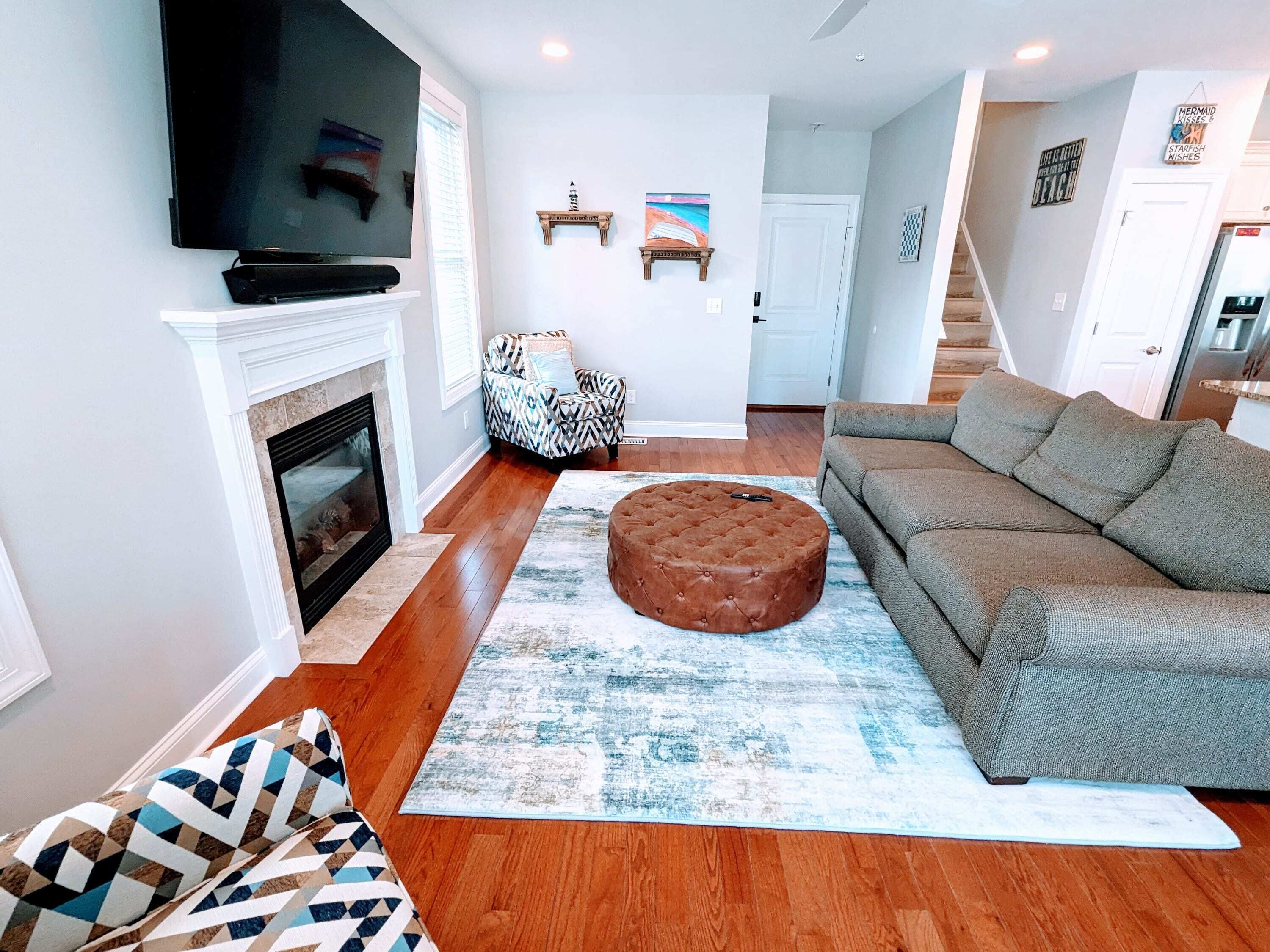 Una sala de estar con chimenea, un televisor montado encima, un sillón estampado, un reposapiés redondo y un sofá gris.