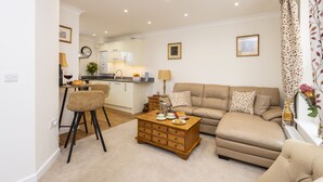 Een woonkamer met een bank, salontafel en stoelen. Er is een keukenhoek met een aanrecht en kastjes.