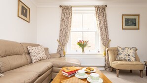 Een gezellige woonkamer met een beige bank, een houten salontafel met boeken, kopjes en een schaakbord, en een raam met gordijnen.