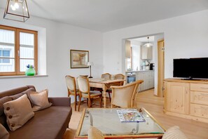 Ett vardagsrum med en soffa, ett soffbord i glas, ett matbord med stolar, en köksdel och en tv.