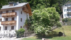 A multi-story building with balconies, a large flowering tree, and a stone wall. - Charming Apartment “Holunder” with Mountain View, Wi-Fi, Balcony & Garden (Bad Tölz)