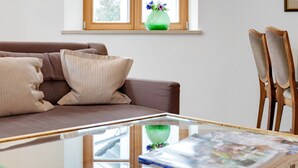 A living room with a brown sofa, a glass coffee table, a wooden chair, and a window with a green vase. - Charming Apartment “Holunder” with Mountain View, Wi-Fi, Balcony & Garden (Bad Tölz)