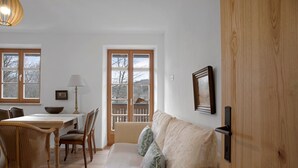 A room with a wooden door, a sofa with cushions, a dining table with chairs, and a window with a view of trees. - Charming Apartment “Ferienwohnung Birke” with Mountain View, Wi-Fi (Bad Tölz)