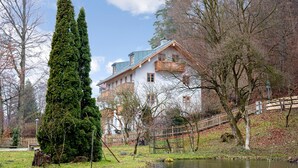 A large building with a sloped roof and multiple windows, surrounded by trees and a small pond. - Charming Apartment “Ferienwohnung Birke” with Mountain View, Wi-Fi (Bad Tölz)