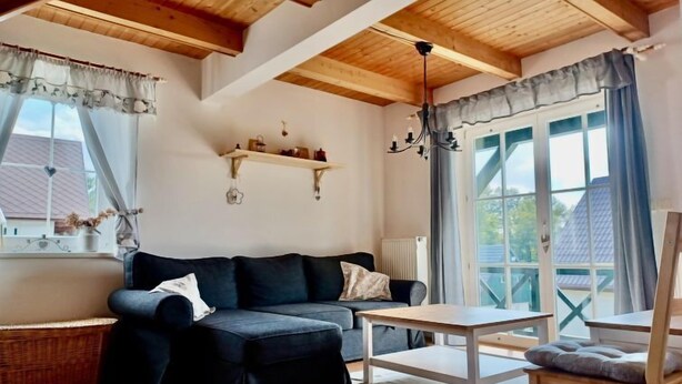 Una sala de estar acogedora con un sofá oscuro, una mesa de centro de madera y una silla. Se observan vigas de madera en el techo y una ventana con cortinas.