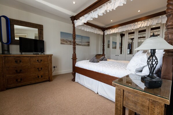 A bedroom with a four-poster bed, a wooden dresser, a flat-screen TV, and a lamp on a glass table.