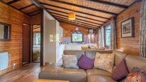 A cozy cabin interior with a leather sofa, wooden walls, and a kitchen area visible through an open doorway.