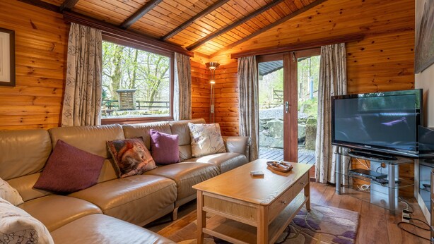 A wooden cabin interior with a leather sofa, a wooden coffee table, a television on a stand, and large windows with curtains.