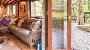 A cozy cabin interior with a leather sofa, wooden beams, and a large window overlooking a forested area.