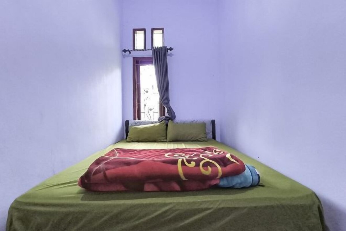 A single bed with a green bedspread and a red blanket, a window with curtains, and a wall with a light blue color.