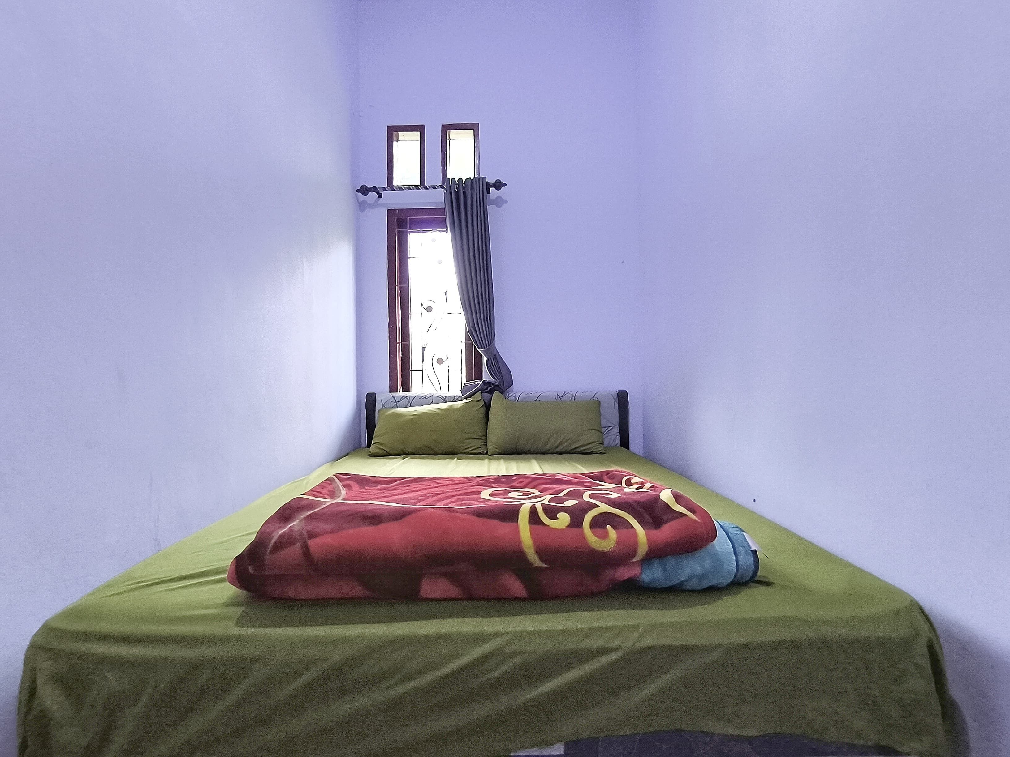A single bed with a green bedspread and a red blanket, a window with curtains, and a door with a wooden frame.