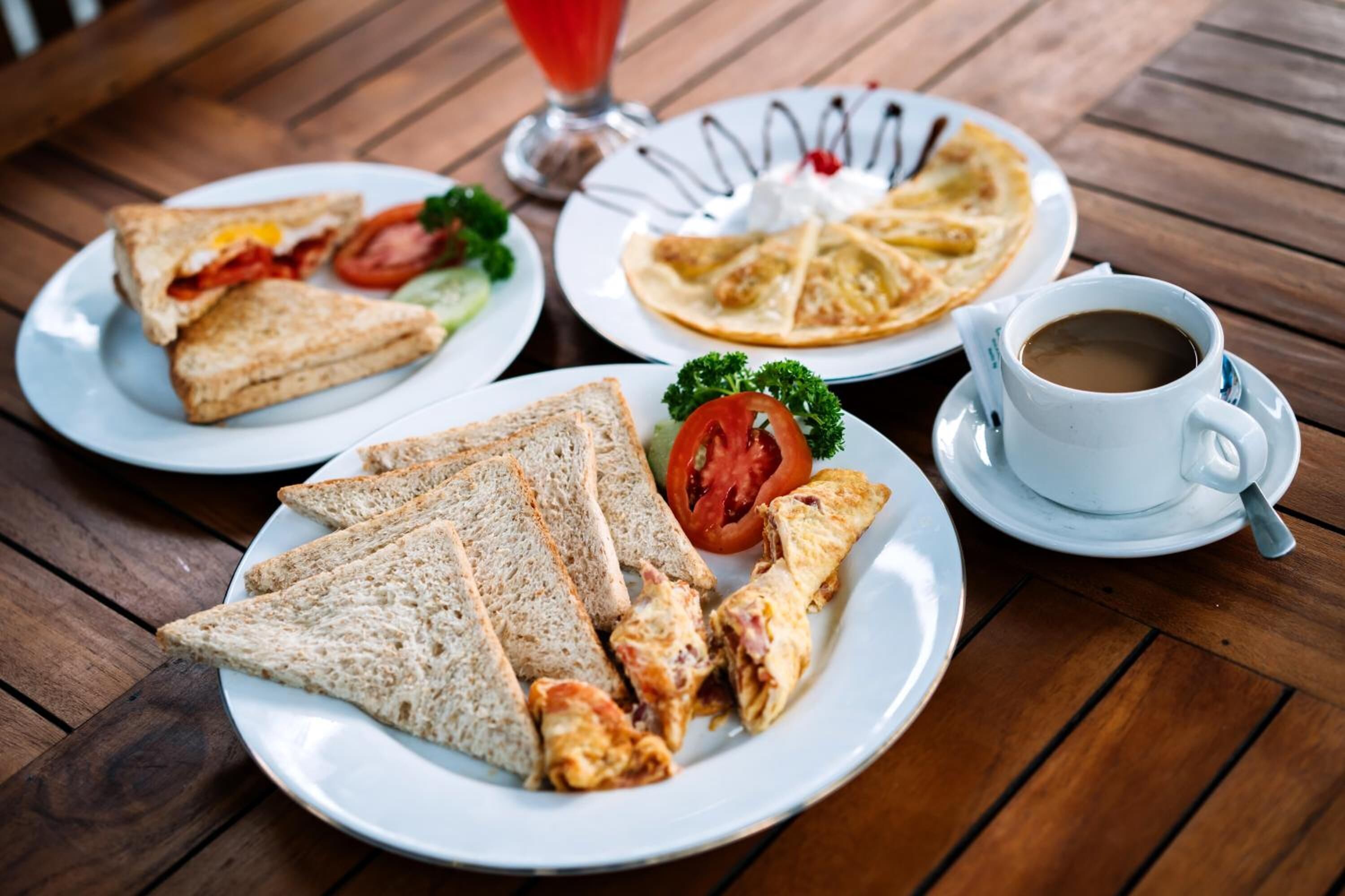 Una mesa de madera con un plato de sándwiches, un plato de panqueques, una taza de café y un vaso de bebida roja.