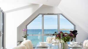 A dining area with a table set for four, large windows with a view of the sea, and a white fluffy chair. - The Penthouse Fistral Beach -  an apartment overlooking Fistral Beach sleeping six (Newquay)