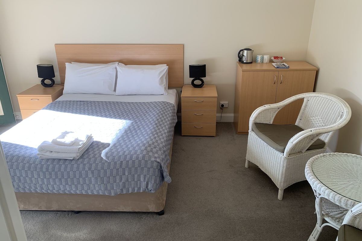A neatly made bed with a checkered duvet, a bedside table with a lamp, a wicker chair, and a small cabinet with a kettle and mugs.