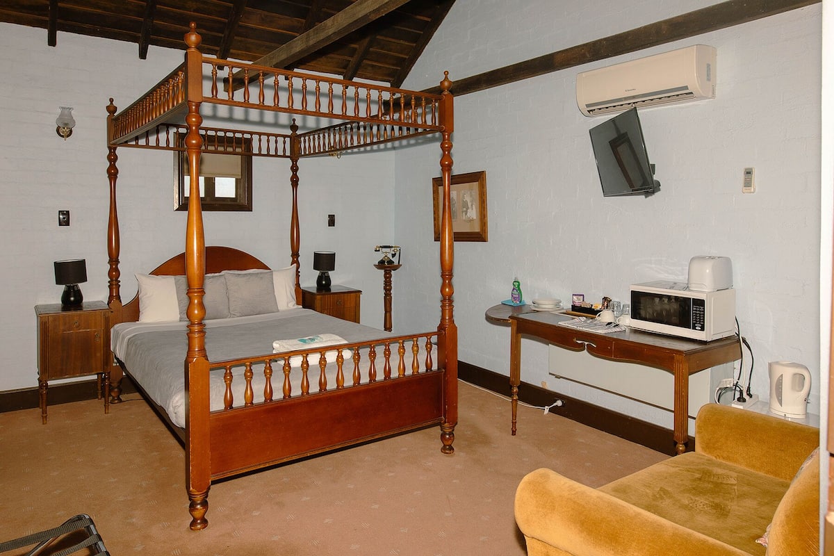 A four-poster bed with a wooden frame, a bedside table with a lamp, a small table with a microwave, and a chair.