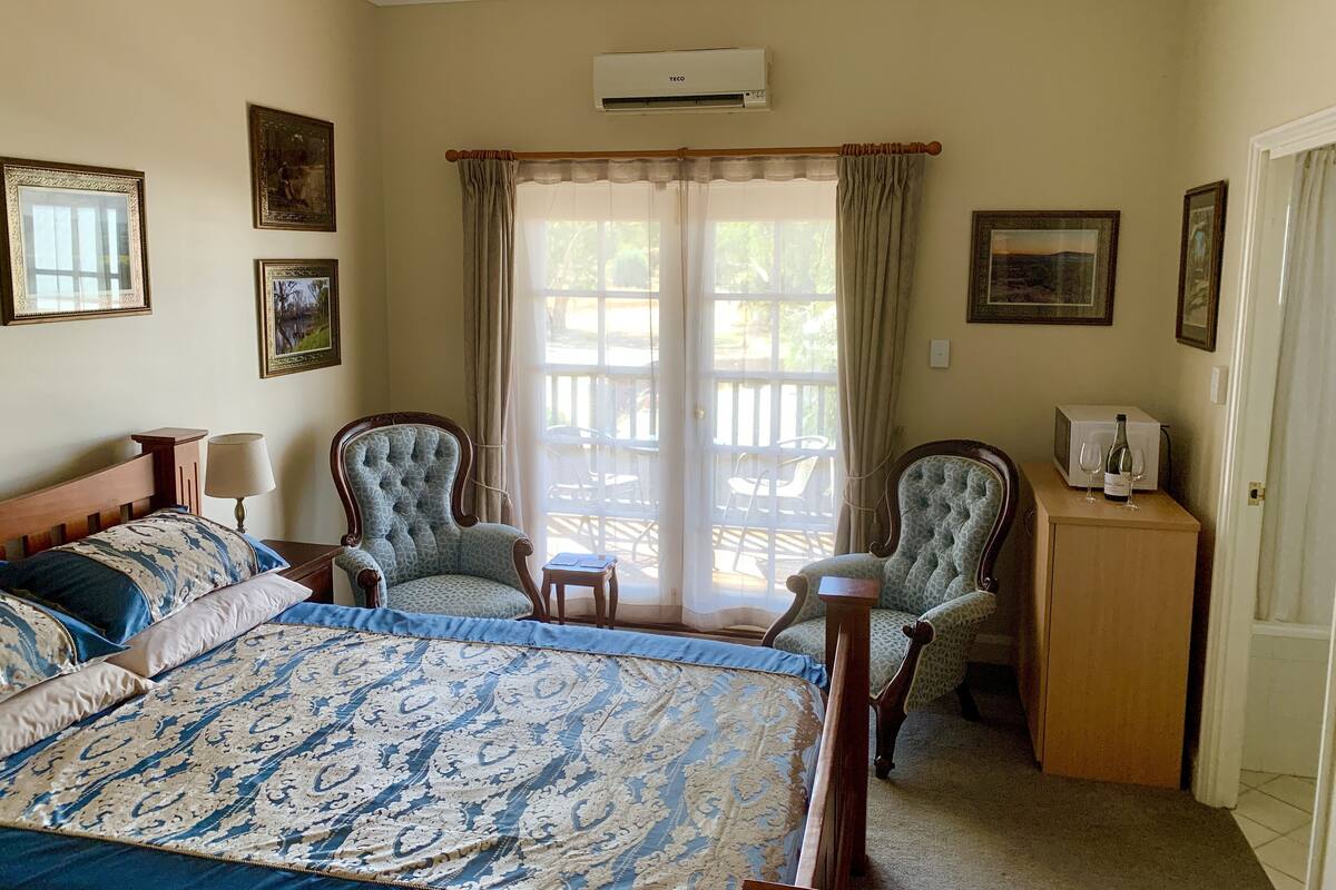A bedroom with a large bed, two armchairs, a wooden nightstand, a TV, and a minibar.