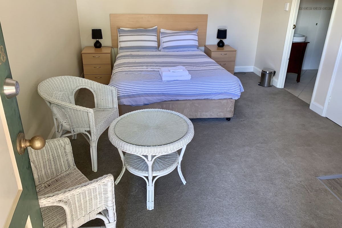 A bedroom with a bed, two nightstands, two wall-mounted lamps, a wicker chair, a round wicker table, and a wooden door.