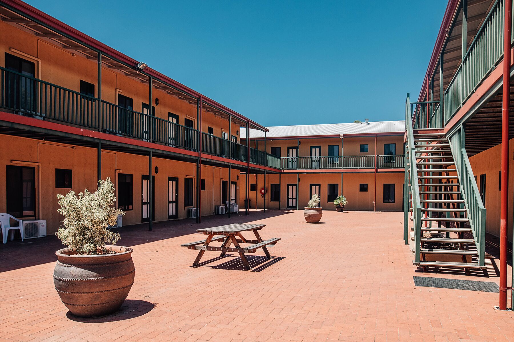 Un patio con una mesa de picnic y plantas en macetas, rodeado de edificios de varios pisos con balcones y una escalera.