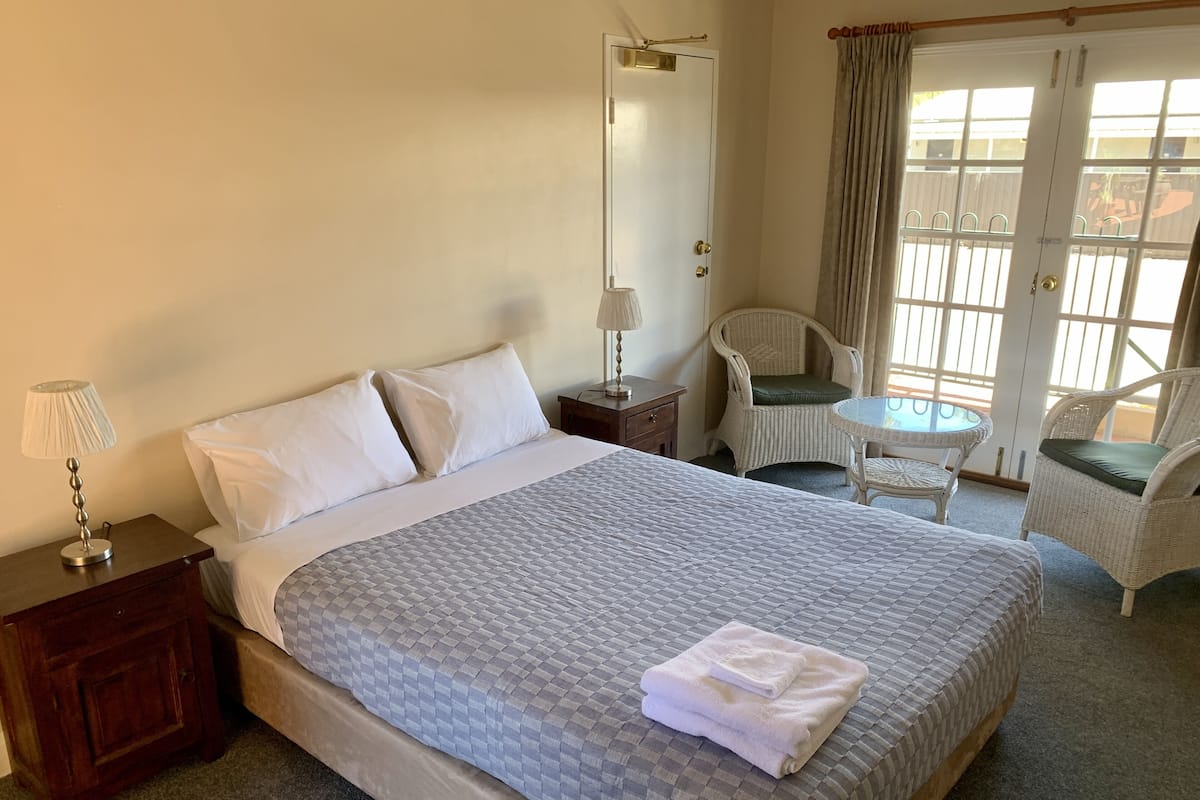 A hotel room with a large bed, a nightstand with a lamp, a chair, a small table, and a view through the window.