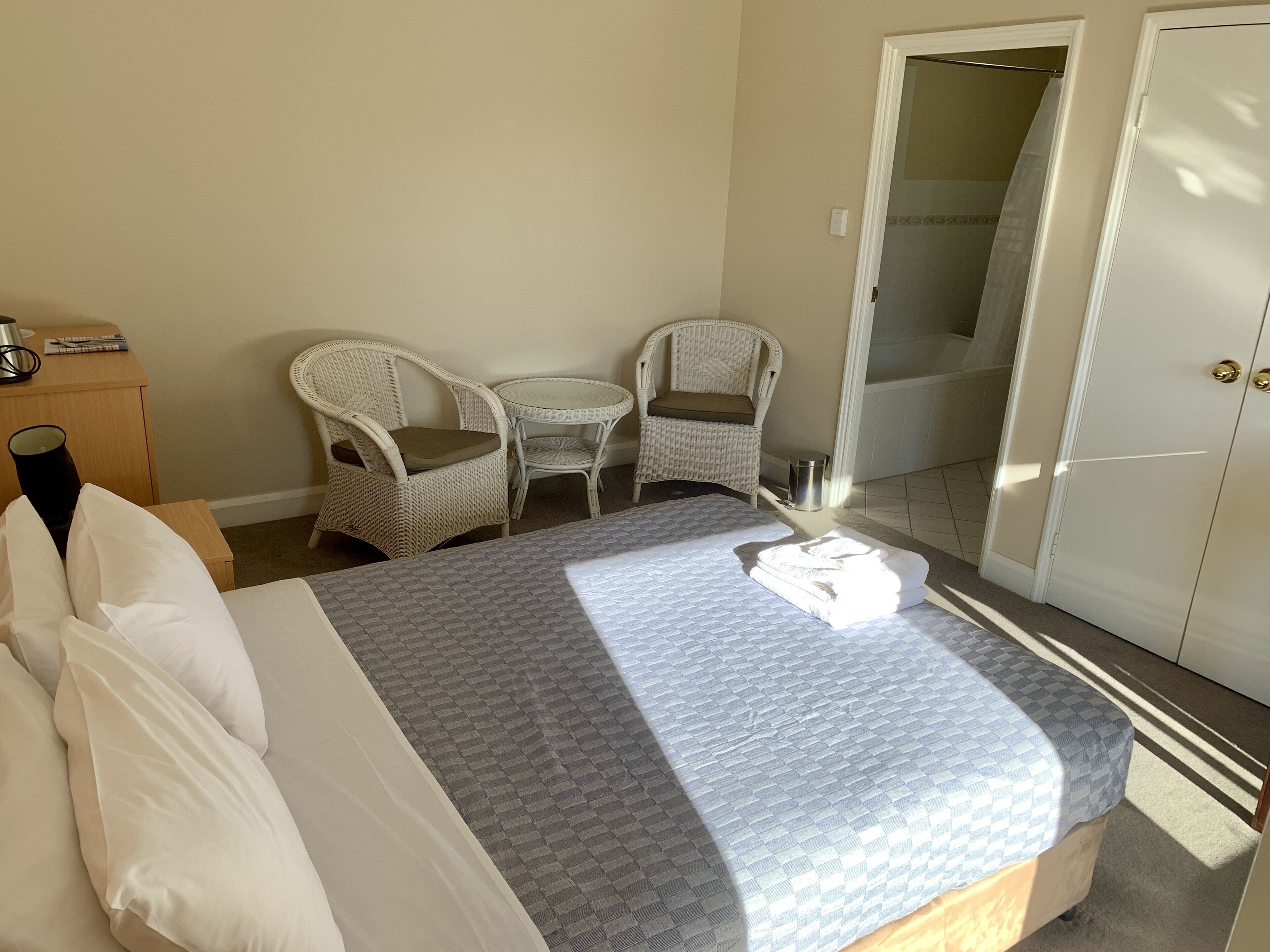 Habitación de hotel con cama, dos sillones de mimbre blanco, una mesita redonda, un armario blanco y zona de ducha.
