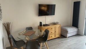 A modern living room with a glass dining table, black chairs, a wooden cabinet, a flat-screen TV, and a window with curtains. - 2 Bedroom River House St Neots (Saint Neots)