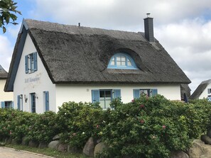 Traditional thatched-roof house with a steeply pitched roof and small dormer window, set amidst a well-maintained garden with pink flowers.