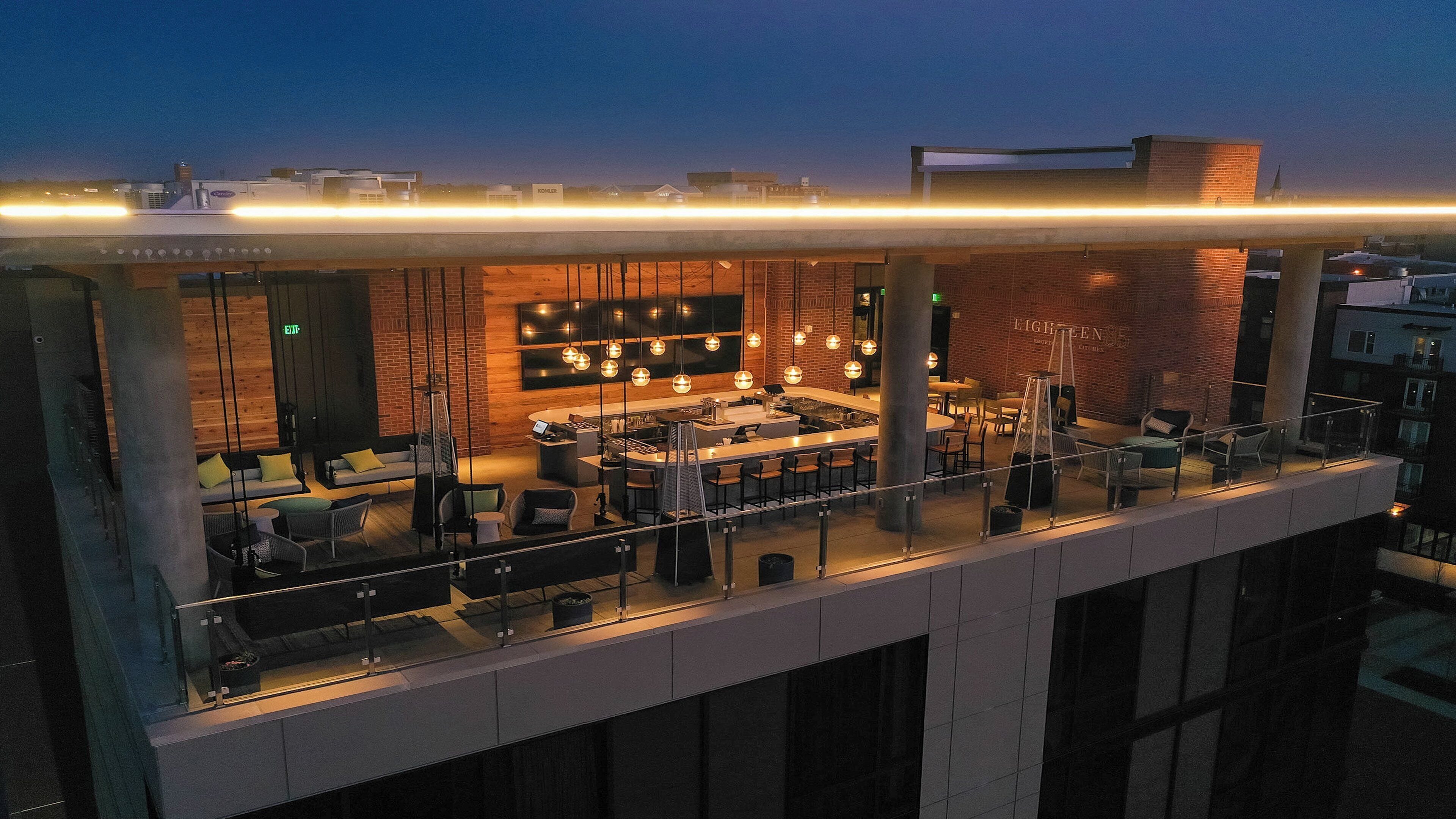 Un bar situé sur le toit d’un immeuble, avec un comptoir, un espace salon et une vue sur les toits de la ville au crépuscule.